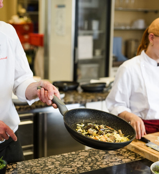 Ein Mann schwenkt Gemüse in einer Pfanne, eine Frau schneidet auf einem Brett. | © Touristikgemeinschaft Hohenlohe e. V. | Wald & Schlosshotel Friedrichsruhe