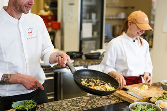 Ein Mann schwenkt Gemüse in einer Pfanne, eine Frau schneidet auf einem Brett. | © Touristikgemeinschaft Hohenlohe e. V. | Wald & Schlosshotel Friedrichsruhe