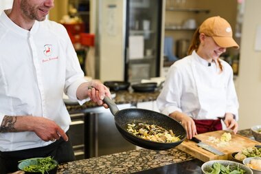 Ein Mann schwenkt Gemüse in einer Pfanne, eine Frau schneidet auf einem Brett. | © Touristikgemeinschaft Hohenlohe e. V. | Wald & Schlosshotel Friedrichsruhe