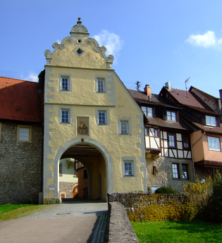 Das Tor ist gelb und hat einen Giebel. Daneben befinden sich Fachwerkhäuser auf der Stadtmauer | © Touristikgemeinschaft Hohenlohe e. V. | Stadt Forchtenberg