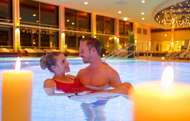 Ein Mann und eine Frau befinden sich im Wasser, der Mann hält die Frau in seinen Armen. Der Innenraum der Therme erstrahlt im Kerchenlicht. | © Björn Hänssler