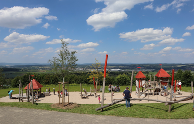 Hölzerne Spielgeräte auf sandigem Boden und Rasenfläche mit Personen