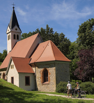 Zwei Radfahrer stehen vor einer Kapelle mit einem spitzen Turm | © Touristikgemeinschaft Hohenlohe e. V. | Susanne Schleussner
