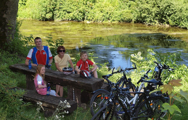 Eine Familie macht bei einer Radtour Rast auf einer Bank an der Jagst | © Touristikgemeinschaft Hohenlohe e. V.  | Susanne Schleussner