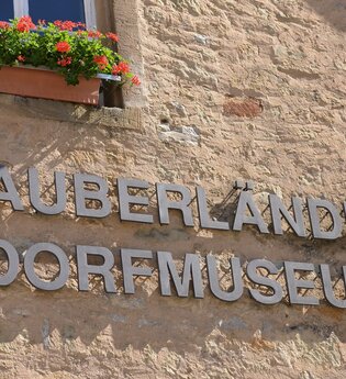 Blick auf den aus einzelnen Metallbuchstaben zusammengesetzen Namen "Tauberländer Dorfmuseum", der auf der Außenseite des Museums angebracht ist.