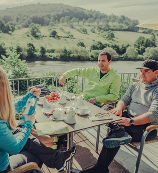 4 Personen sitzen an einem Tisch bei Kaffee und Kuchen; im Hintergrund fließt der Neckar. | © Touristikgemeinschaft Odenwald e.V.
