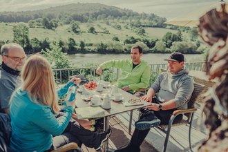 4 Personen sitzen an einem Tisch bei Kaffee und Kuchen; im Hintergrund fließt der Neckar. | © Touristikgemeinschaft Odenwald e.V.