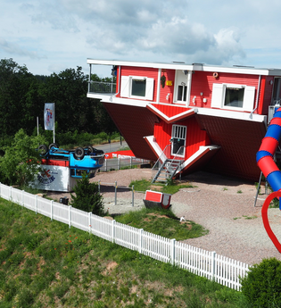 Blick auf das Toppels-Haus "Verdrehte Welt". Das Haus und ein Auto davor stehen auf  dem Kopf. Im Außenbereich ist ein Spielplatz mit Spielgeräten, z.T. auch auf dem Kopf stehend, zu sehen. | © TOPPELS - Verdrehte Welt