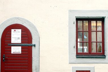 Eingangstüre zur Touristinformation aus dunkelrot gestrichenem Holz, rechts daneben zwei, dunkelrot gestrichene Sprossenfenstertüre, | © Petra Natzkowski