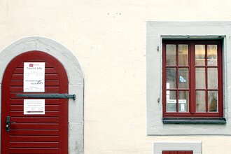 Eingangstüre zur Touristinformation aus dunkelrot gestrichenem Holz, rechts daneben zwei, dunkelrot gestrichene Sprossenfenstertüre, | © Petra Natzkowski