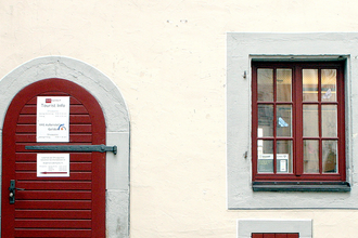 Eingangstüre zur Touristinformation aus dunkelrot gestrichenem Holz, rechts daneben zwei, dunkelrot gestrichene Sprossenfenstertüre, | © Petra Natzkowski