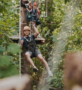 Person mit Helm und Kletterausrüstung schwebt an einer Seilrutsche im Wald, weitere Personen warten auf einer Plattform im Hintergrund | © TG HeilbronnerLand / Tourismus im Weinsberger Tal | Niklas Hesser