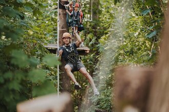 Person mit Helm und Kletterausrüstung schwebt an einer Seilrutsche im Wald, weitere Personen warten auf einer Plattform im Hintergrund | © TG HeilbronnerLand / Tourismus im Weinsberger Tal | Niklas Hesser