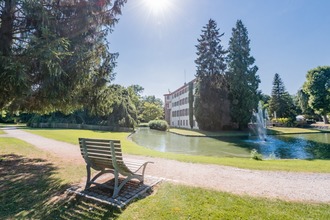 Schlosspark mit Teich, Brunnen und Schloss unter blauem Himmel