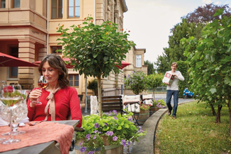 In der linken Hälfte sitzt eine Frau mit einem Weinglas in der Hand an einem gedeckten Tisch. In der rechten Hälfte läuft eine Person auf einer Gras bewachsenen Fläche, zwischen kleinen Bäumen, mit einem Weinkarton in der Hand.