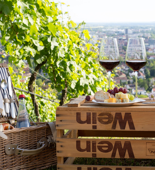 links ist ein geöffneter, mit blau-weiß gestreiften Stoff ausgekleideter Picknickkorb befüllt mit Backwaren und Wasser zu sehen daneben auf einer umgedrehten Weinkiste befindet sich ein Teller mit Käse und Weintrauben und zwei mit Rotwein befüllte Gläser | © Heilbronn Marketing GmbH