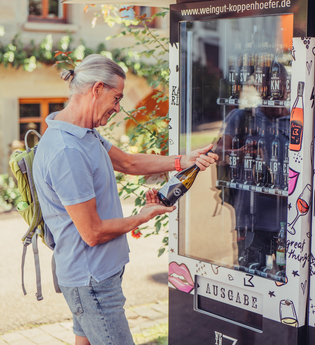 Ein Mann steht am Weinautomaten vom Weingut Koppenhöfer und hält eine Weinflasche in der Hand | © Tourismus im Weinsberger Tal e.V.