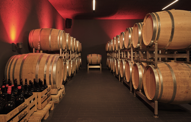 Dunkler Raum beleuchtet durch wenige gedimmte Deckenfluter. Rechts und links im Bild sind Weinfässer zu erkennen, jeweils zwei Reihen liegend übereinander. Im Bild rechts vorne stehen zwei, mit Weinflaschen gefüllte Weinkisten | © Weingut Albrecht-Kiessling