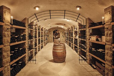 eleganter Gewölbekeller mit mehreren, gut bestückten, Weinregalen rechts und links. In der Mitte des Kellers steht ein Weinfass | © Weingut Albrecht-Kiessling
