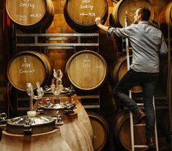 Winzer Becker auf der Leiter vor seinen beschrifteten hölzernen Weinfässern im Weinkeller. | © Weingut Becker in Malsch
