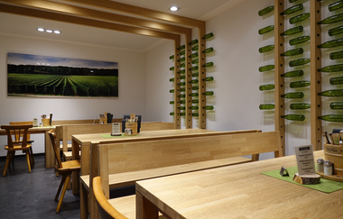 heller Raum mit drei langen Tischen aus Holz, Weinflaschen in Konstruktion an der Wand | © Christian Seyffer