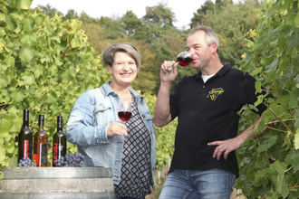 Eine Frau und ein Mann stehen in einem Weinberg bei einem Weinfass und halten ein Glas Rotwein in der Hand | © Touristikgemeinschaft Hohenlohe e. V. | Weinhof Dieroff