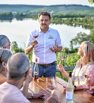 Ein gemischte Gruppe bekommt einen Wein erklärt. Der Weinausschank liegt oberhalb vom Breitenauer See, der im Hintergrund zu erkennen ist. | © Touristikgemeinschaft HeilbronnerLand