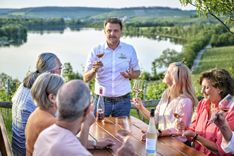 Ein gemischte Gruppe bekommt einen Wein erklärt. Der Weinausschank liegt oberhalb vom Breitenauer See, der im Hintergrund zu erkennen ist. | © Touristikgemeinschaft HeilbronnerLand