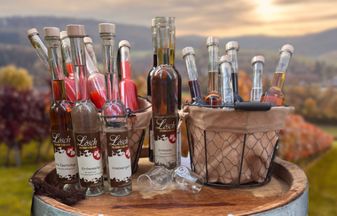 Verschiedene Brände und Liköre, schön auf einem Weinfass präsentiert, mit Sonnenuntergang im Hintergrund | © Touristikgemeinschaft Hohenlohe e. V. | Winzerstube Lösch