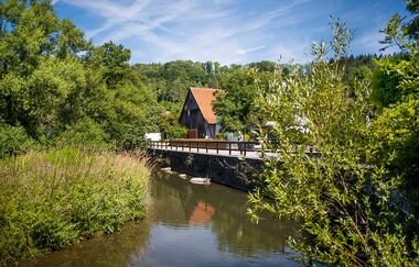 Blick auf die Bühler (Gewässer), über welches eine Brücke führt. An der Brücke steht ein Haus, welches von Bäumen und Natur umgeben ist. | © Nicole Hagemann & Marco Emminger (@kommwirmachendaseinfach)
