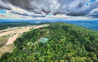 Drohnenaufnahme auf Berg mit kleinem See. Der See ist umgeben von einem grünen Wald. Am Horizont sind Hügel zu erkennen. Der Himmel ist hellblau mit vielen Wolken. | © TMBW, Foto: Dietmar Denger