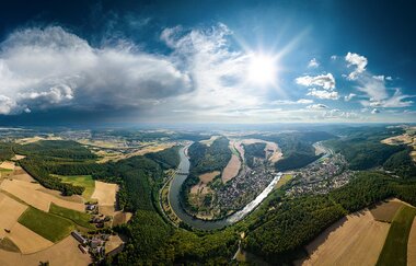 Drohnenaufnahme des Naturparks Neckartal-Odenwald. Der Himmel ist strahlend Blau mit Wolken, die Sonne scheint direkt in die Kamera. Landschaftlich ist eine Neckarschleife zu erkenne, umgeben von Wald und Feldern. | © TMBW, Foto: Dietmar Denger