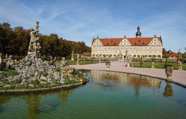 Im Vordergrund ist ein großer Brunnen, welcher mit Wasser befüllt ist. In der Mitte des Brunnen ist eine Statue als Wasserfontäne. Im Hintergrund steht imposant das Schloss Weikersheim. Der Himmel ist strahlend blau mit wenigen Wolken. | © Susanne Maier