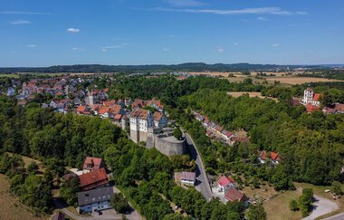 Blick auf Vellberg von oben. Das Schloss Vellberg ist zentral im Bild, umgeben von der Stadt, vielen Bäumen und Feldern. Am Horizont ist eine Hügellandschaft. Der Himmel ist strahlend blau mit wenigen Wolken. | © Nicole Hagemann & Marco Emminger (@kommwirmachendaseinfach)