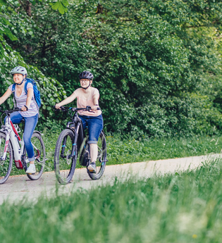 2 Radfahrerinnen fahren auf einem Radweg mit ihren E-Bikes. Der Radweg führt durch Wiesen und Wälder. | © Touristikgemeinschaft Odenwald e.V.