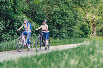 2 Radfahrerinnen fahren auf einem Radweg mit ihren E-Bikes. Der Radweg führt durch Wiesen und Wälder. | © Touristikgemeinschaft Odenwald e.V.