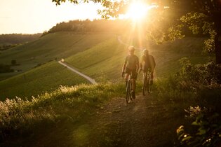Mann und Frau gravelbiken über die Höhenzüge des Kraichgau-Strombergs. Im Hintergrund Sonnenuntergang.