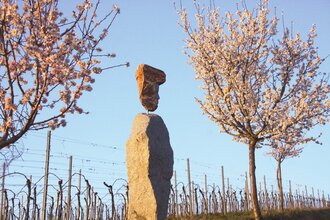 Eine Steinskulpur aus zwei Felsbrocken, stellen einen Weingott dar, im Hintegrund sind Weinreben zu sehen, links und rechts stehen blühende Mandelbäume. | © Maria Zimmermann