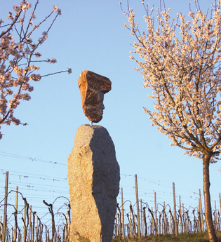 Eine Steinskulpur aus zwei Felsbrocken, stellen einen Weingott dar, im Hintegrund sind Weinreben zu sehen, links und rechts stehen blühende Mandelbäume. | © Maria Zimmermann