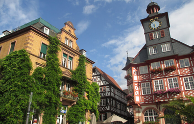 Links steht ein gelbes Sandsteingebäude mit grünem Bewuchs, rechts steht das Fachwerkbau des Rathauses mit großem Glockenturm. | © Maria Zimmermann