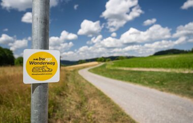 Ein "bwegt-Wanderweg"-Schild befindet sich auf einem Pfosten an einem Feldweg.