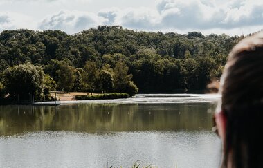 Eine Frau blickt auf den See, im Hintergrund sind Bäume und Liegefläche zu sehen.