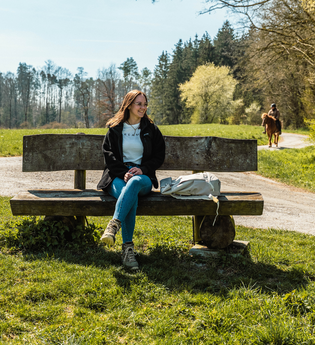 Eine junge Frau sitzt auf einer Bank auf einer Wiese, die von Wald umgeben ist. | © Stadtverwaltung Crailsheim