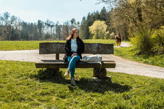 Eine junge Frau sitzt auf einer Bank auf einer Wiese, die von Wald umgeben ist. | © Stadtverwaltung Crailsheim