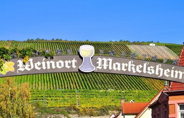 Ein Schild mit der Aufschrift "Weinort Markelsheim", dekoriert mit einem Weinglas. Im Hintergrund sieht man die Weinberge. | © Thomas Weller