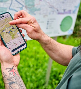 Eine Person prüft auf dem Smartphone den Verlauf eines Wanderwegs; im Hintergrund steht eine große Wanderkarte. | © Tourimia Tourismus GmbH