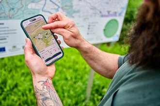 Eine Person prüft auf dem Smartphone den Verlauf eines Wanderwegs; im Hintergrund steht eine große Wanderkarte. | © Tourimia Tourismus GmbH