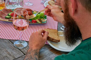 Ein Mann mit Bart und Tattoos genießt eine deftige Vesper mit Wurst, Käse, Trauben und Brot bei einem Glas Roséwein auf kariertem Tischtuch. | © Kraichgau-Stromberg Tourismus e.V.
