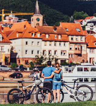 2 Radler stehen mit Ihren E-Bikes am Neckarufer und betrachten die Altstadt von Eberbach, ein Schiff fährt vorbei. | © Touristikgemeinschaft Odenwald e.V.