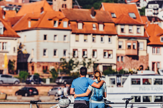 2 Radler stehen mit Ihren E-Bikes am Neckarufer und betrachten die Altstadt von Eberbach, ein Schiff fährt vorbei. | © Touristikgemeinschaft Odenwald e.V.